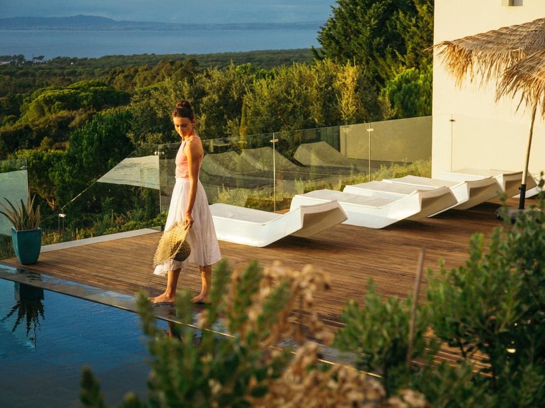 A woman in a white dress walking along the wooden deck by the pool, with sun loungers beside her and the forest and the sea beyond