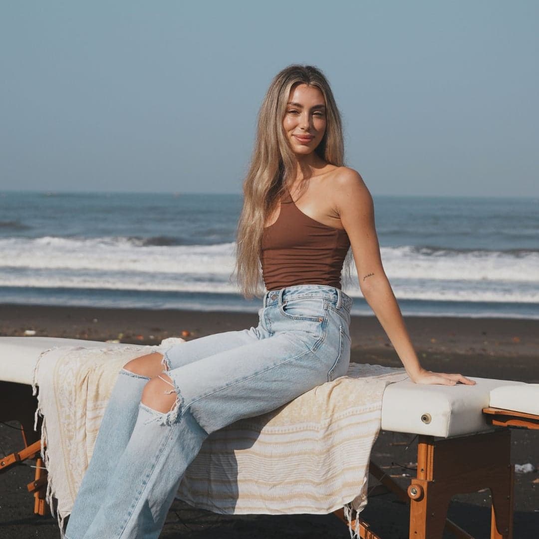 Julia sitting on a treatment table on a black sand beach, the sea behind her