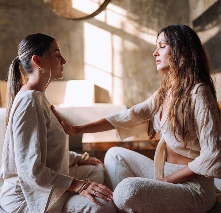 Two women sitting face to face, one resting a hand on the other’s shoulder